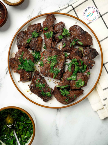 Grilled or seared beef heart slices topped with fresh chopped parsley herbs on a white plate, served next to a bowl of green chimichurri sauce.