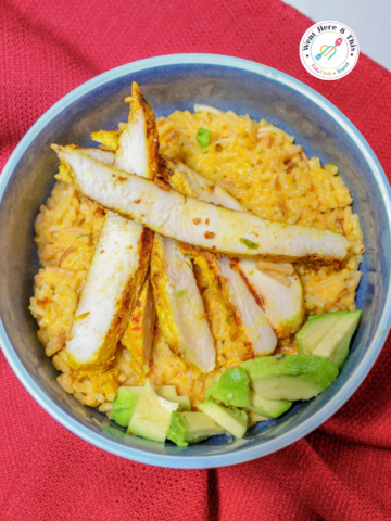 A blue ceramic bowl filled with the Best Lemongrass Chicken Recipe. Slices of juicy, seasoned golden chicken breast are arranged on a bed of flavorful yellow rice mixed with vermicelli noodles. Fresh chunks of green avocado sit on the side. The dish rests on a textured red cloth background under a white circular website logo.
