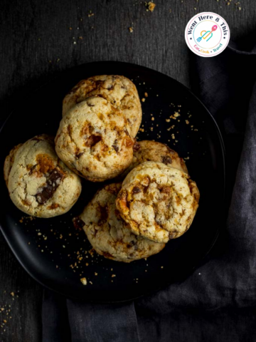 A stack of thick, chewy golden-brown cookies packed with melted chocolate chunks and crushed orange Butterfinger candy pieces, arranged on a matte black plate with a dark grey napkin.