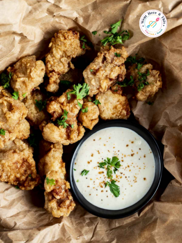 op-down view of golden brown, crispy fried alligator bites garnished with fresh chopped parsley on brown crinkled paper next to a small black bowl filled with creamy dipping sauce.