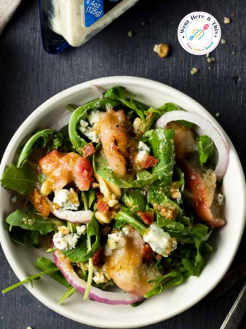 Top-down view of a fresh grilled peach salad in a white bowl with baby arugula, sliced red onions, chopped walnuts, crumbled blue cheese, and a warm bacon vinaigrette drizzle.