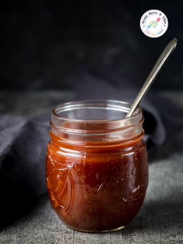Thick glossy homemade barbecue sauce filled inside a glass Ball mason jar with a silver spoon, placed against a dark moody background.