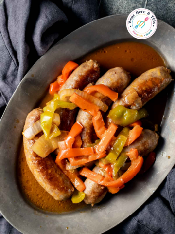 Over-the-shoulder view of cooked Italian sausage links on a rustic metal platter, topped with sliced red and green bell peppers and sweet onions in a rich savory sauce