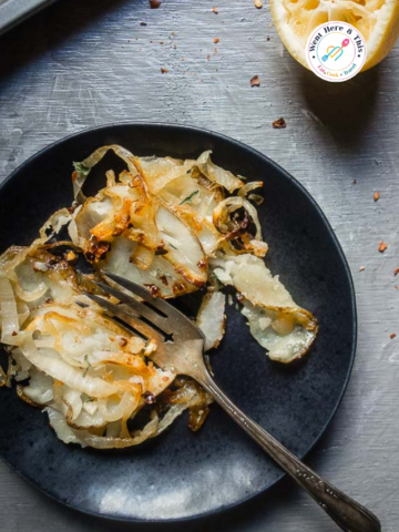 Oven baked Lyonnaise potatoes on a black plate with caramelized onions, garlic, and fresh herbs, served next to a halved lemon on a rustic grey background.