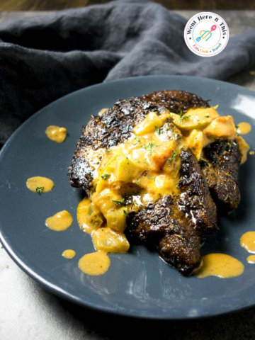 A close-up shot of pan-fried Cajun blackened catfish fillets on a dark gray plate, topped with a rich, creamy yellow Creole sauce and fresh parsley flakes.