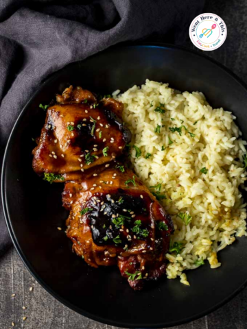 Top-down view of slow cooker teriyaki chicken thighs with a sticky glaze, sesame seeds, and parsley, served in a black bowl alongside seasoned white rice.