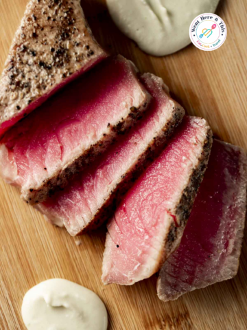 Vertical close-up view of sliced medium-rare sous vide tuna steak with a seared pepper crust on a wooden cutting board with two dollops of wasabi cream sauce.
