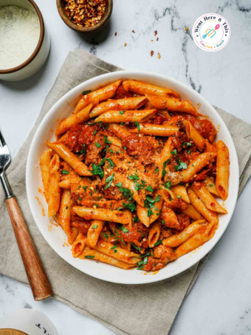 Overhead view of a white ceramic bowl filled with spicy chicken penne pasta tossed in a vibrant red tomato harissa sauce, heavily topped with grated parmesan cheese and fresh chopped parsley on a grey marble tabletop.