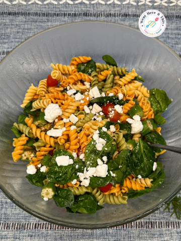 Vibrant summer okra pasta salad made with tri-color rotini pasta, sliced sautéed okra, fresh baby spinach leaves, halved cherry tomatoes, and crumbled feta cheese served in a large clear glass bowl.