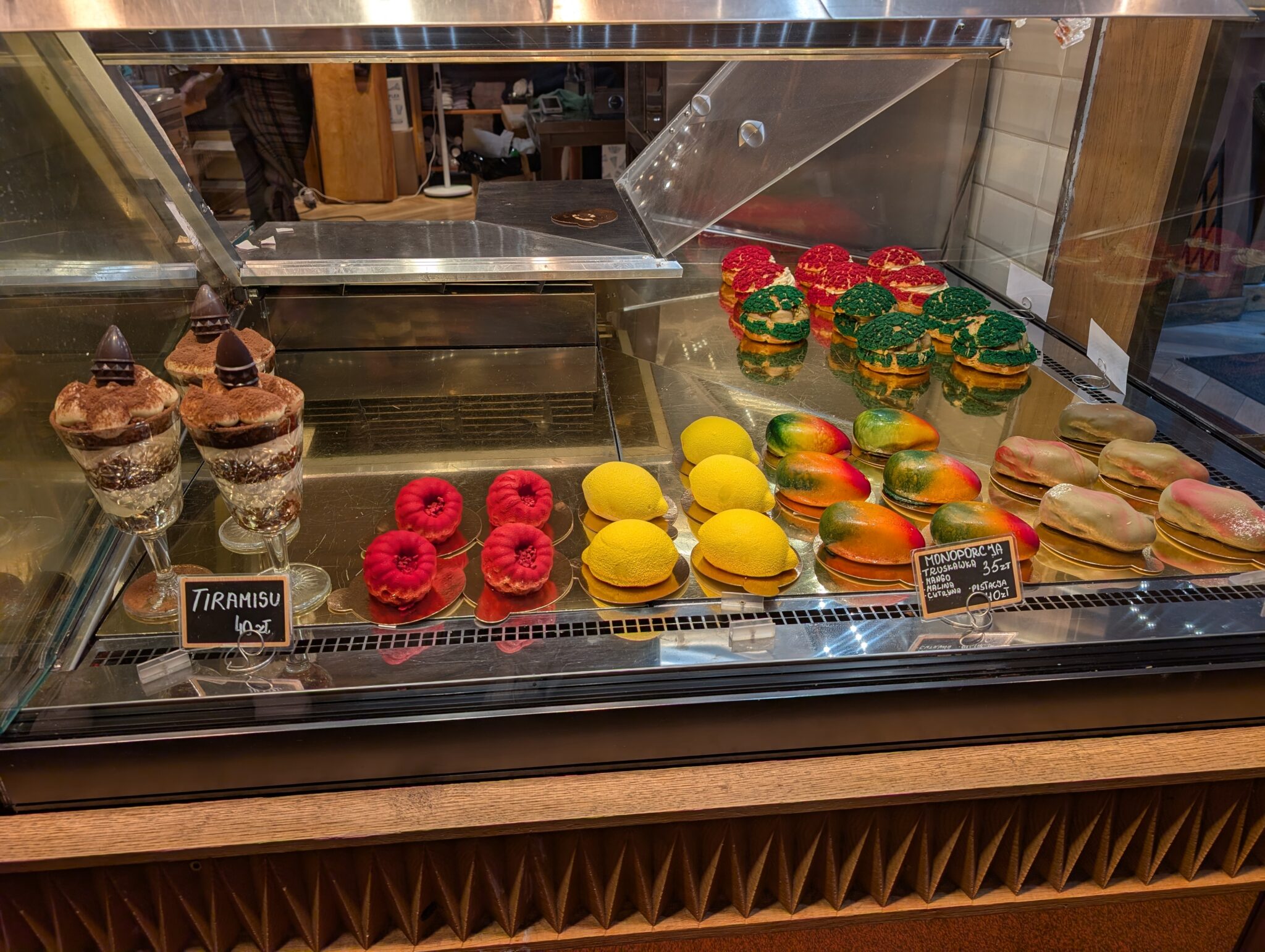 The display case features an assortment of artisanal sculpted fruit-shaped mousse pastries, including hyper-realistic mango, lemon, and raspberry designs alongside tiramisu cups.
