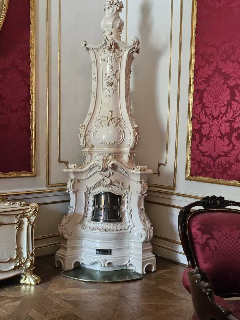 This ornate, multi-tiered white ceramic tile stove (Kachelofen) in the Rococo or Baroque revival style, is typically found in 18th to 19th-century Central European palaces and historic mansions. This particular stove is located in the Sisi Museum.