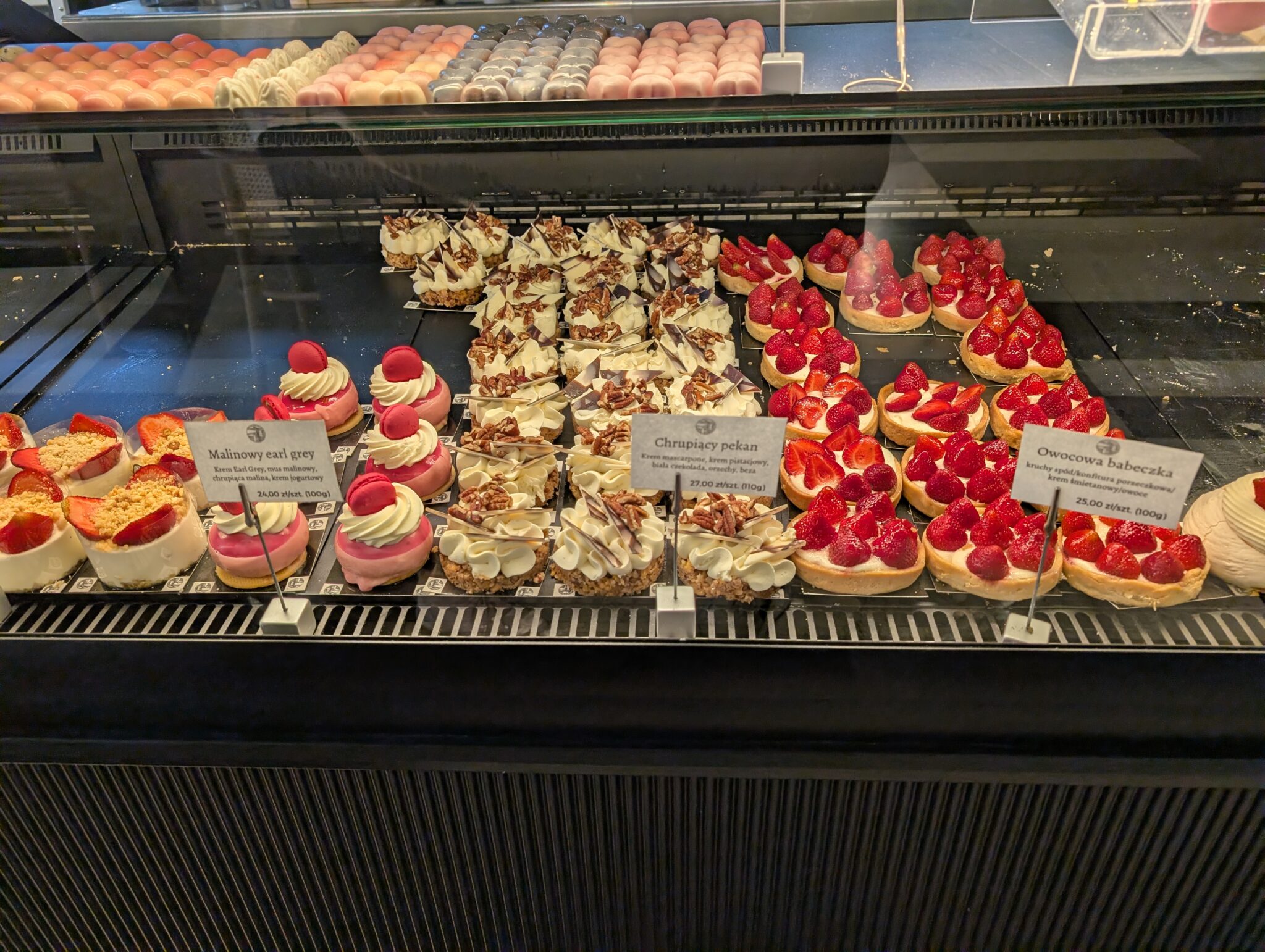 Display case featureing an assortment of fine pastries and tarts, with varieties such as "Malinowy earl grey" and "ChrupiÄ…cy pekan".
