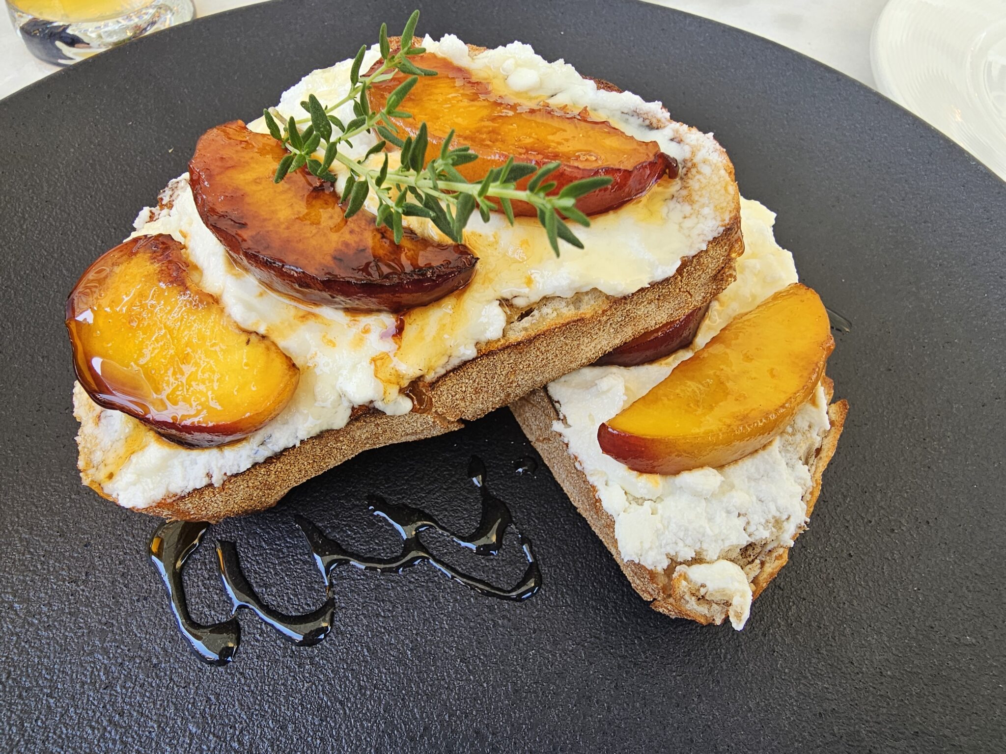 Artisanal toast with creamy mascarpone cheese, caramelized fresh peaches, a drizzle of honey, and a fresh thyme sprig.