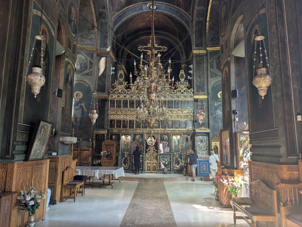 The interior of the historic Curtea Veche Church (also known as the Church of St. Anthony) in Bucharest. The interior features traditional Eastern Orthodox mural paintings, dark wood paneling, and an ornate gilded iconostasis at the front holding sacred icons.