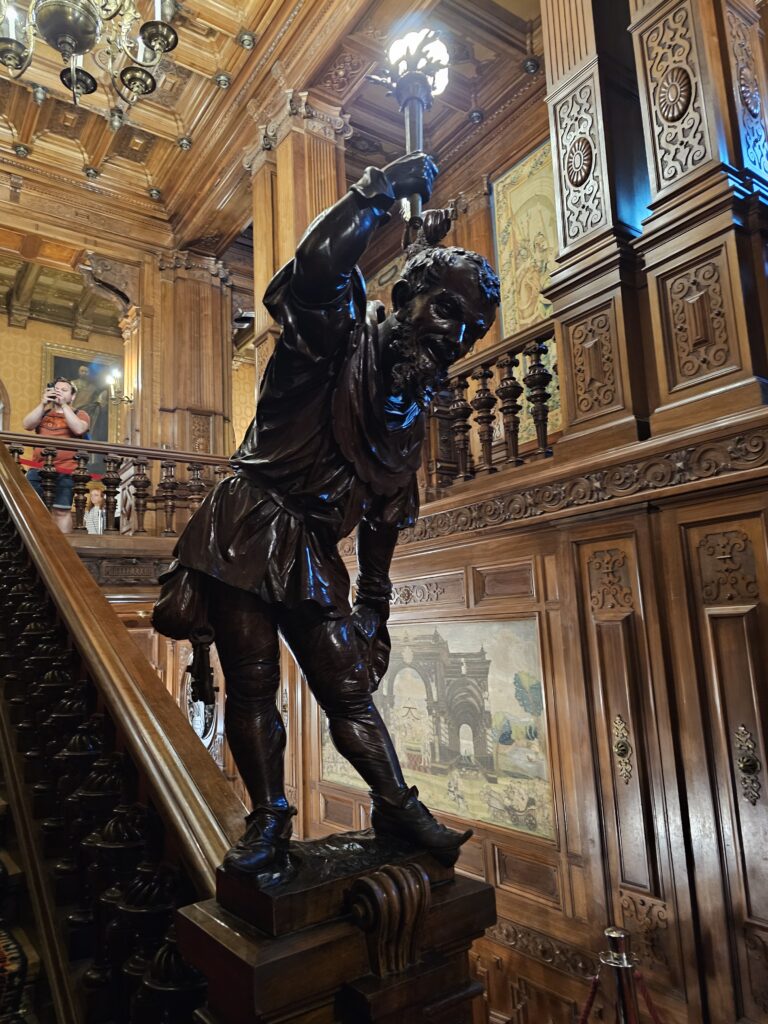 A beautifully carved dark wooden statue holding a torch lamp stands on the main staircase inside PeleÈ™ Castle